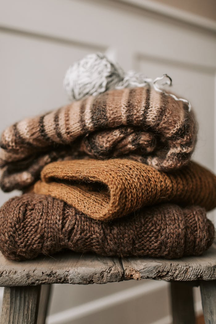 Stack of warm knitted sweaters in earthy tones on a rustic wooden stool indoors.